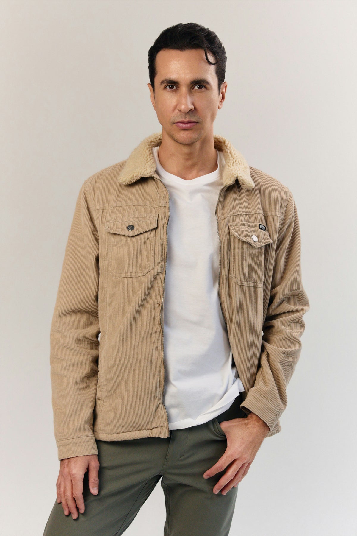 A man wearing a beige corduroy utility jacket with a sherpa collar, layered over a white t-shirt and olive green pants, standing against a light gray studio background.